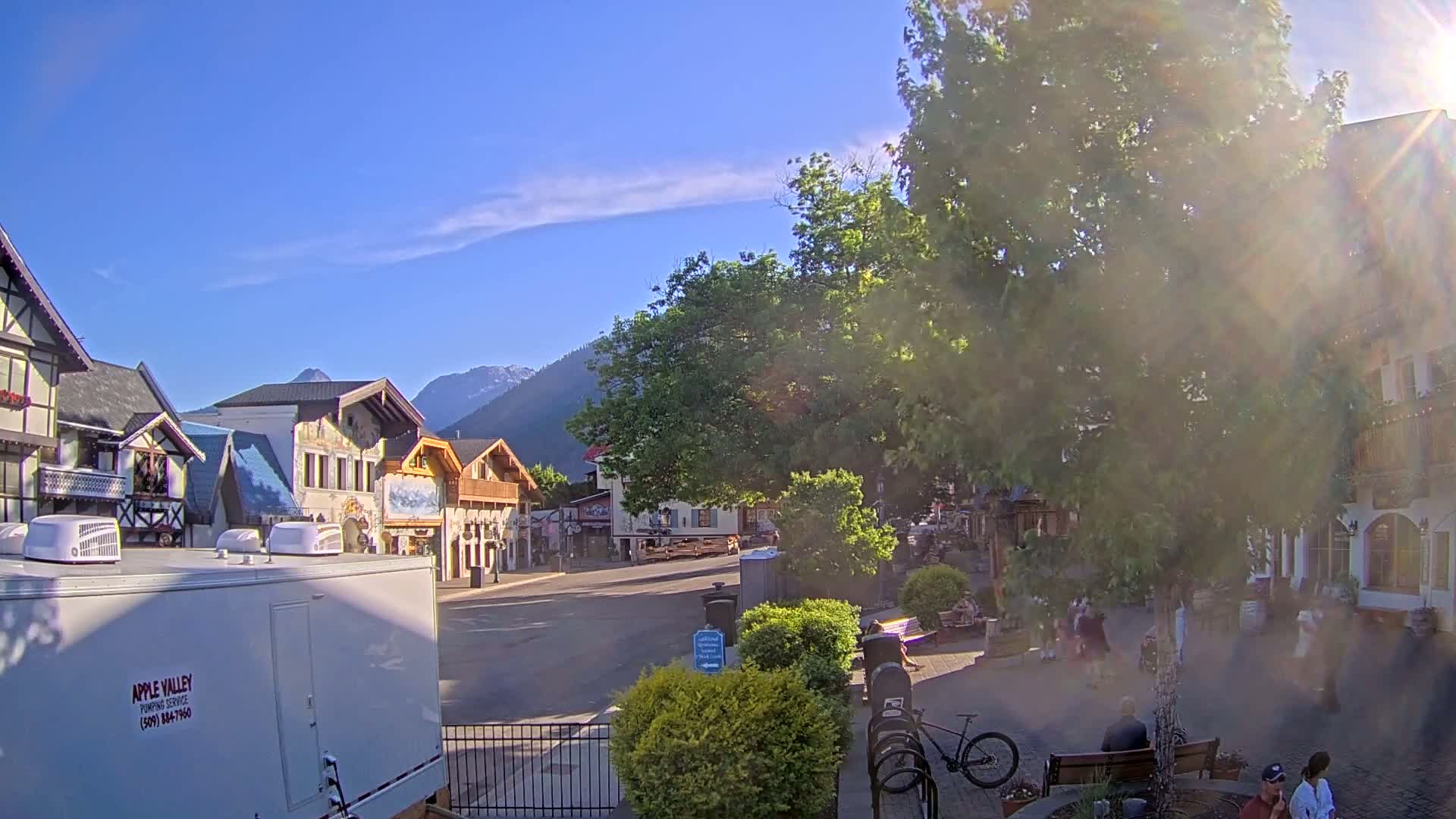 A sunny day in a village square with European-style buildings, a large tree in the center, and several people sitting on benches.