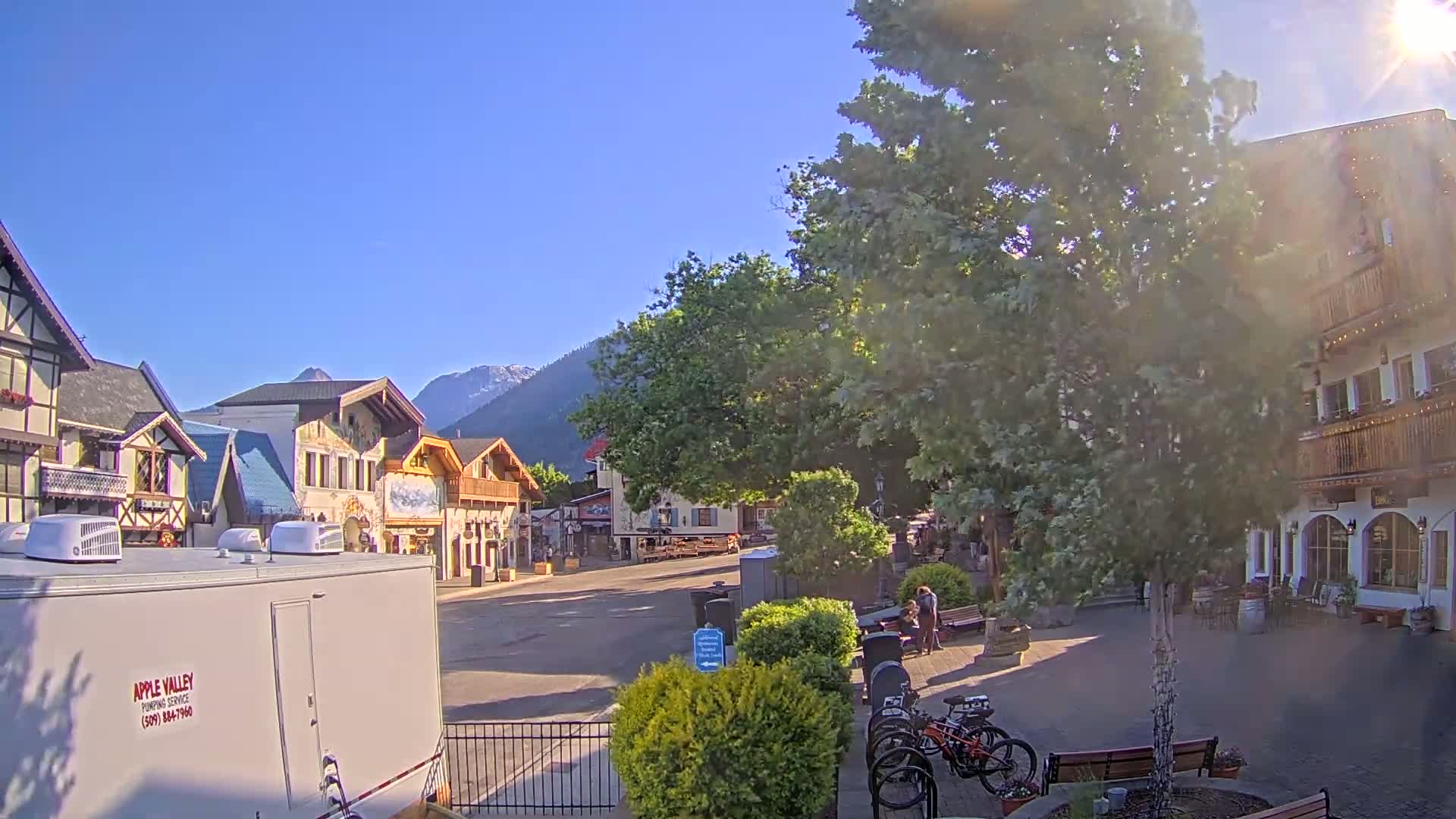 A sunny day reveals a village square with several multi-storied buildings in an alpine style, some bicycles parked nearby, and mountains in the background.