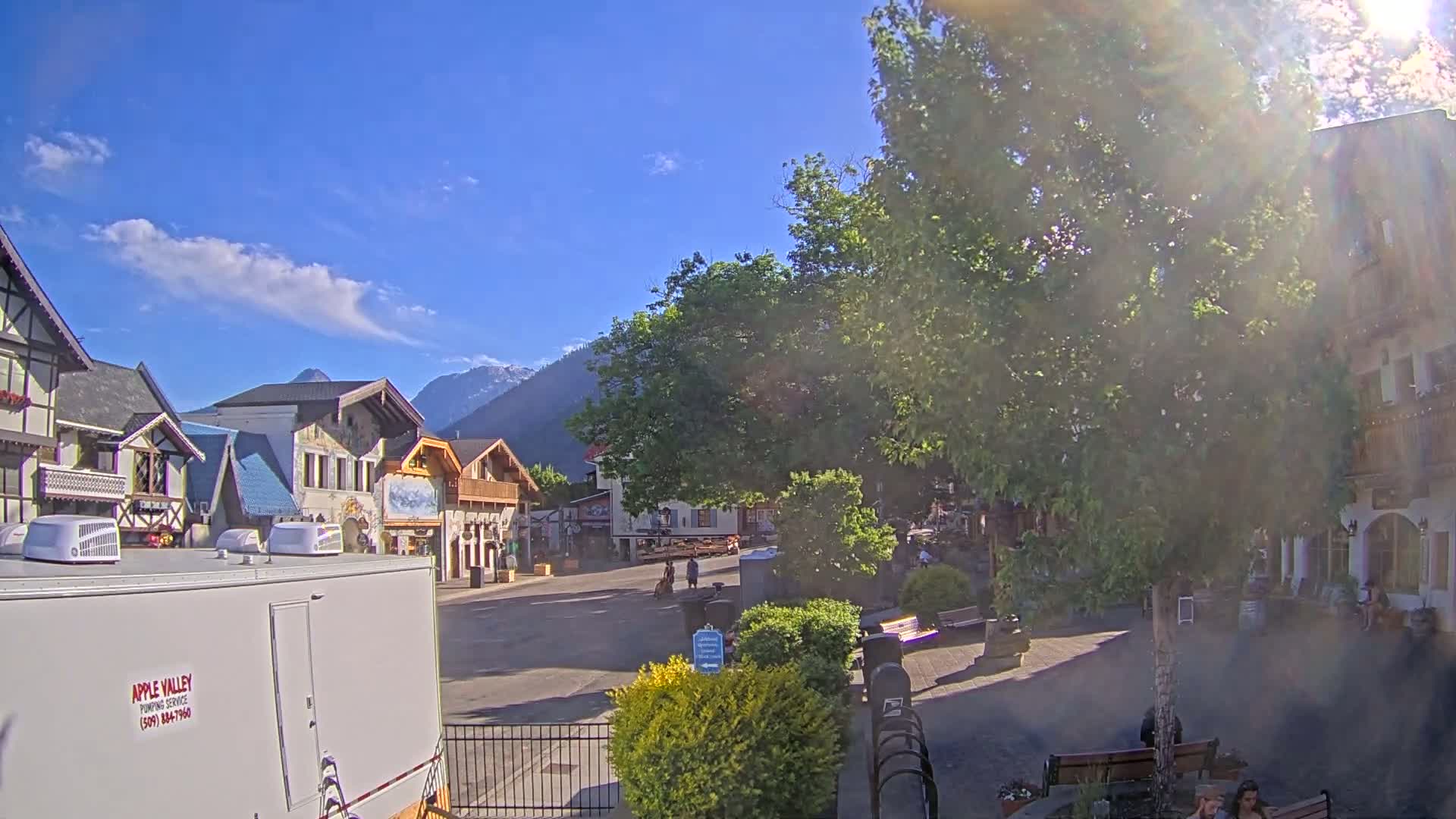 A sunny day in a village square with European-style buildings, a large tree in the center, and mountains in the background.
