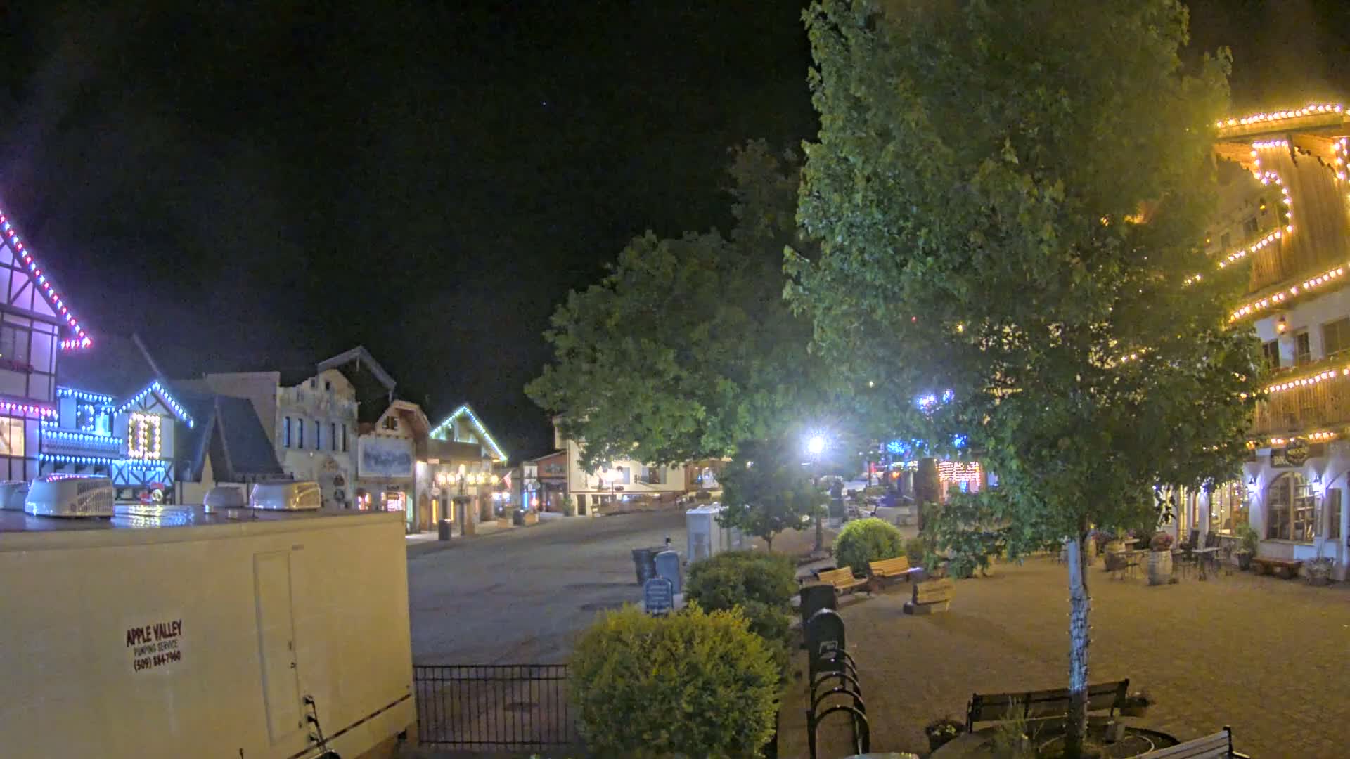 A nighttime view of a town square with several buildings decorated with string lights, a large tree in the center, and benches.