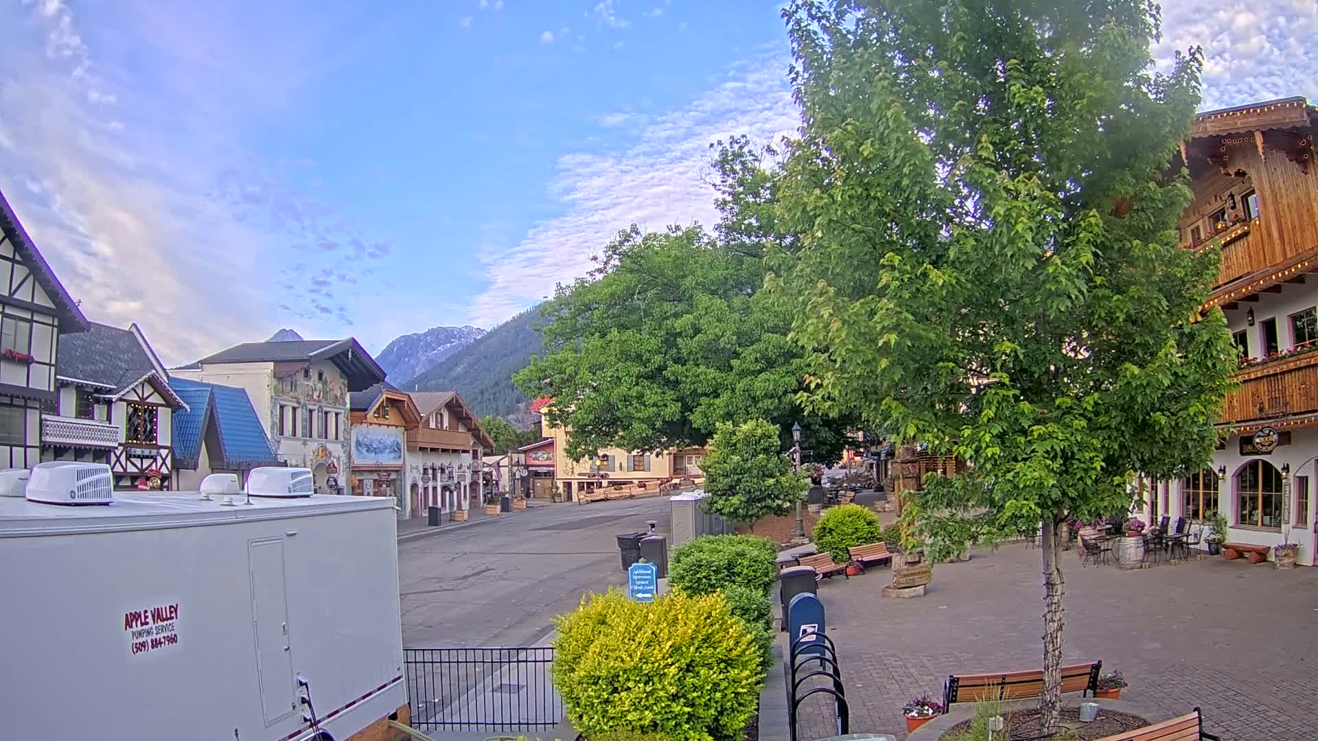 A mostly sunny day reveals a quaint village square with European-style buildings, lush green trees, and mountains in the background.