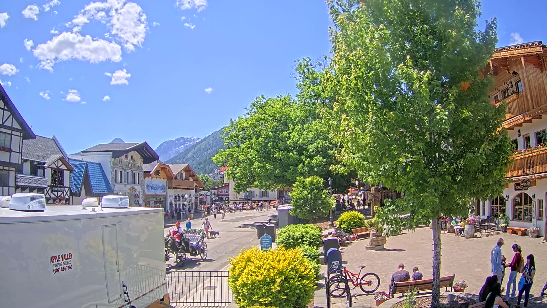 A sunny day in a village square with European-style buildings, a horse-drawn carriage, people walking and sitting, and mountains in the background.