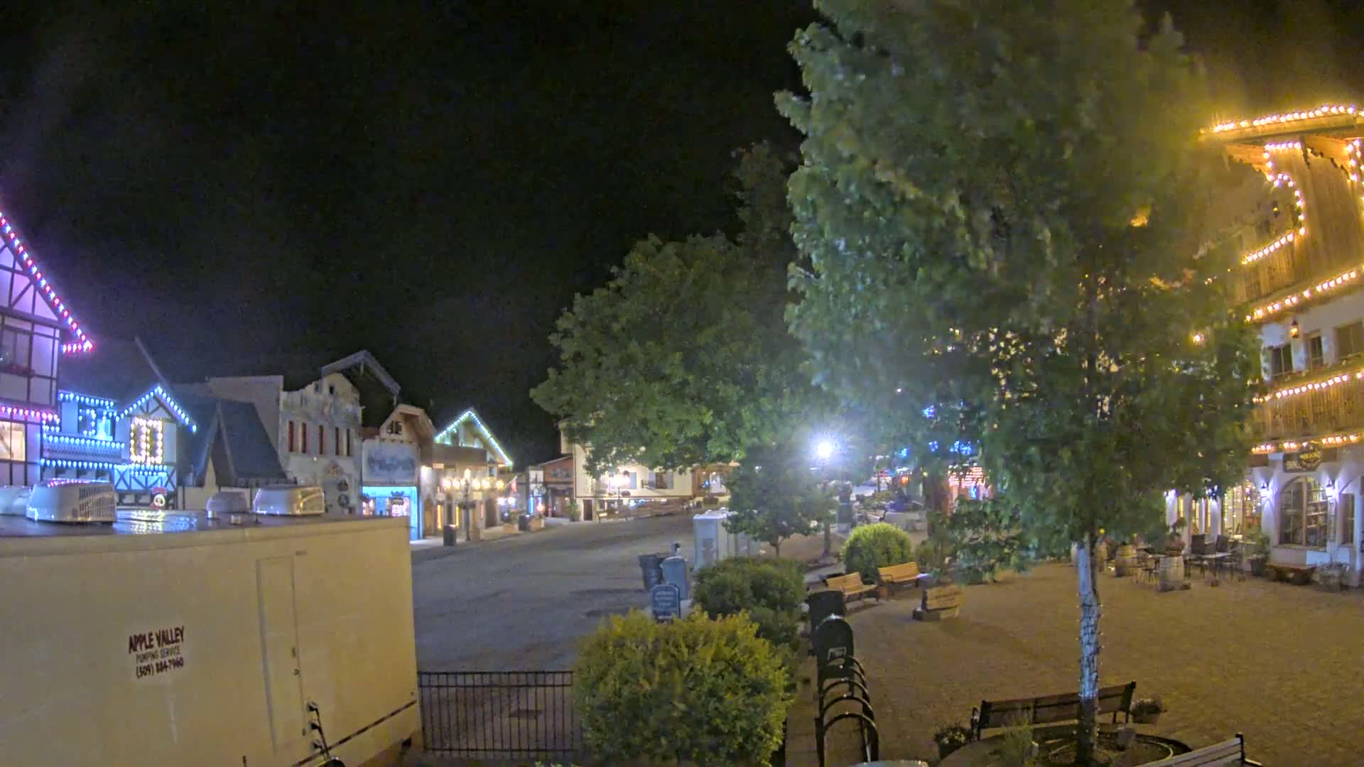 A nighttime view of a town square with several buildings decorated with string lights, trees, and park benches under a dark sky.