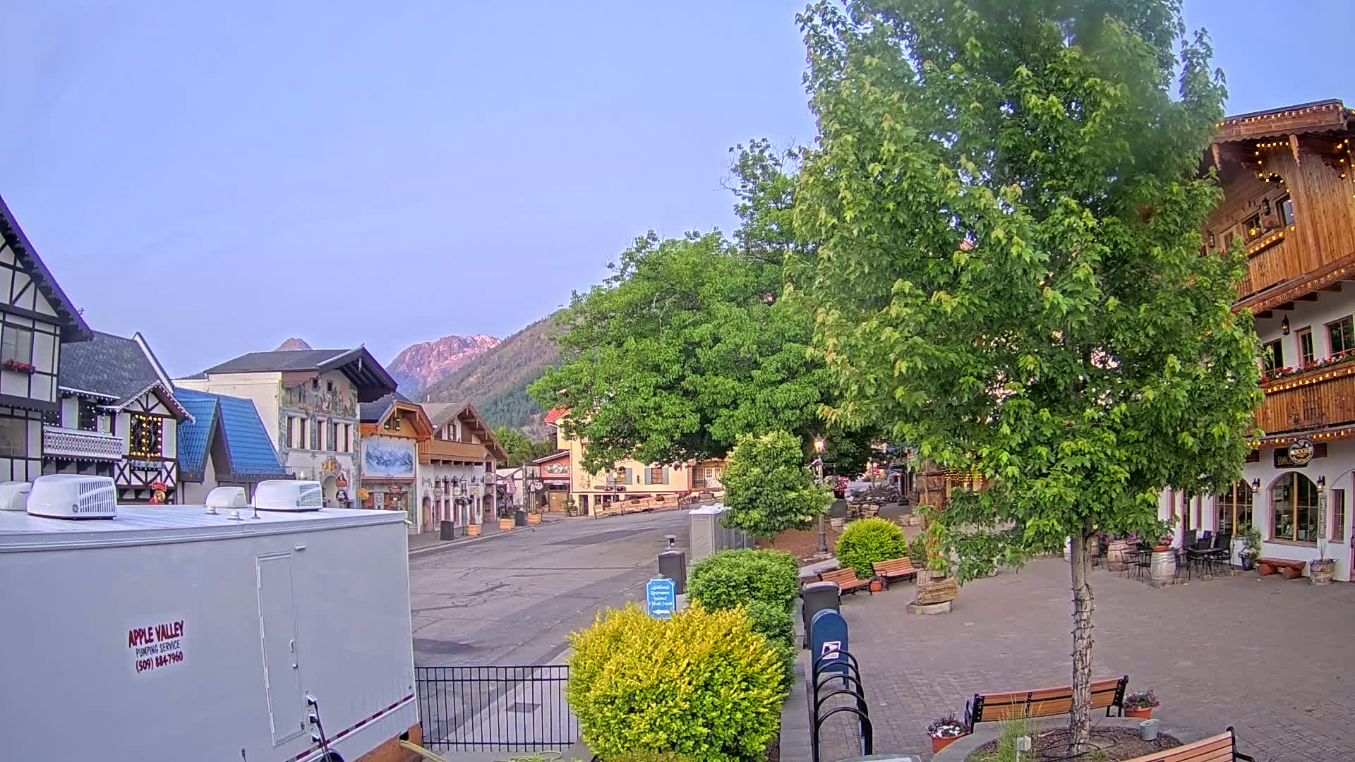 A mostly empty town square with Bavarian-style buildings, lush green trees, and mountains in the background under a clear, light-blue sky.