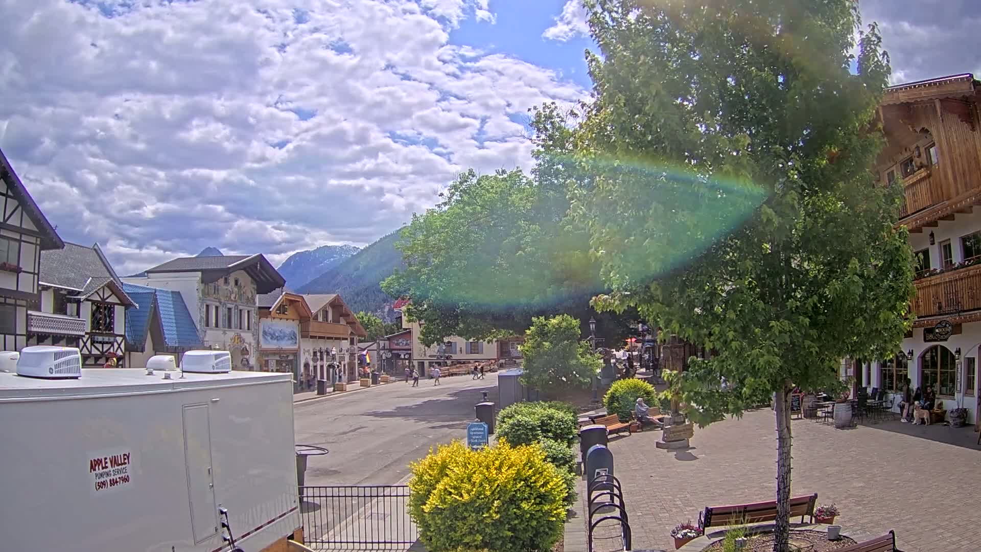 A partly sunny day in a European-style village square with several people walking and sitting, surrounded by buildings and trees, mountains in the background.