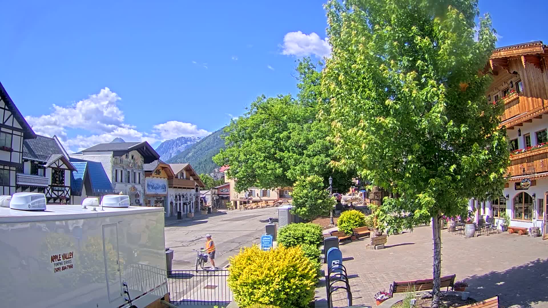 A sunny day in a village square with several buildings, a person riding a bicycle, and lush green trees.