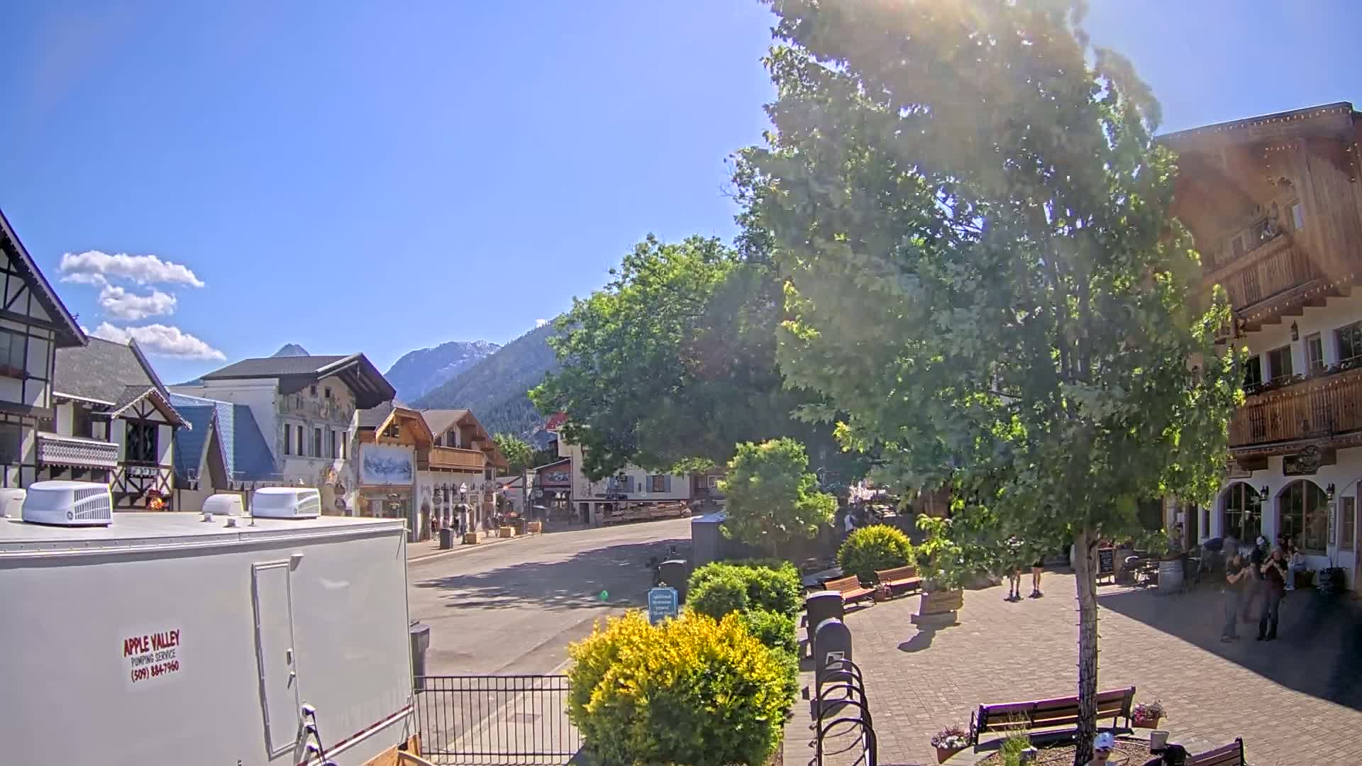 A sunny day in a village with European-style buildings, a paved town square, and lush green trees.