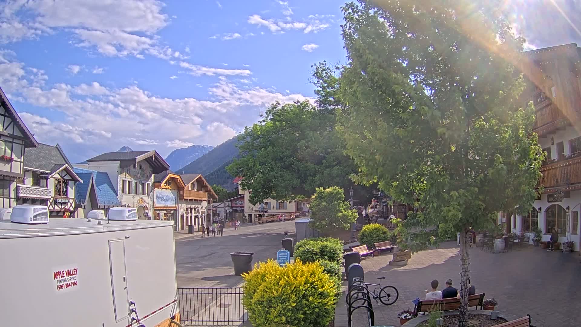 A sunny day in a village square with several European-style buildings, a large tree, some people sitting on a bench, and mountains in the background.
