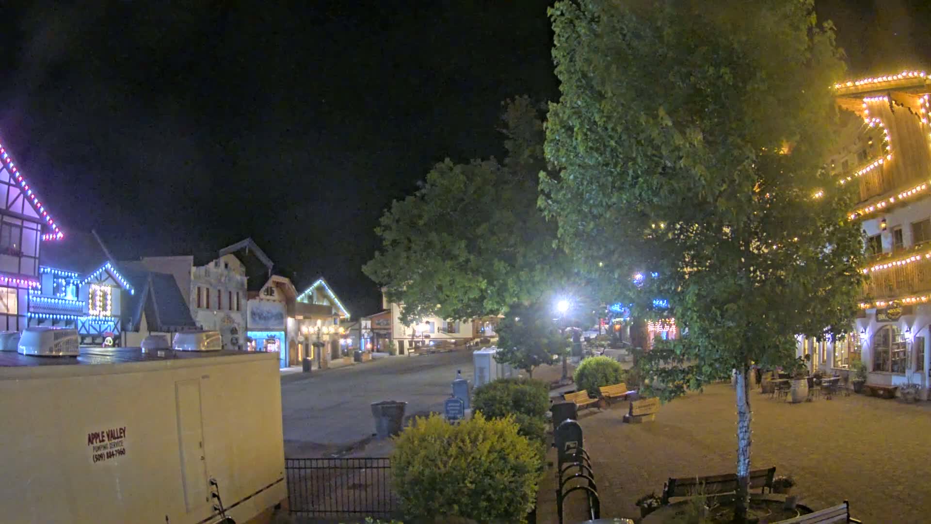 At night, a town square with fairy lights strung along buildings and trees is mostly empty except for some outdoor seating.