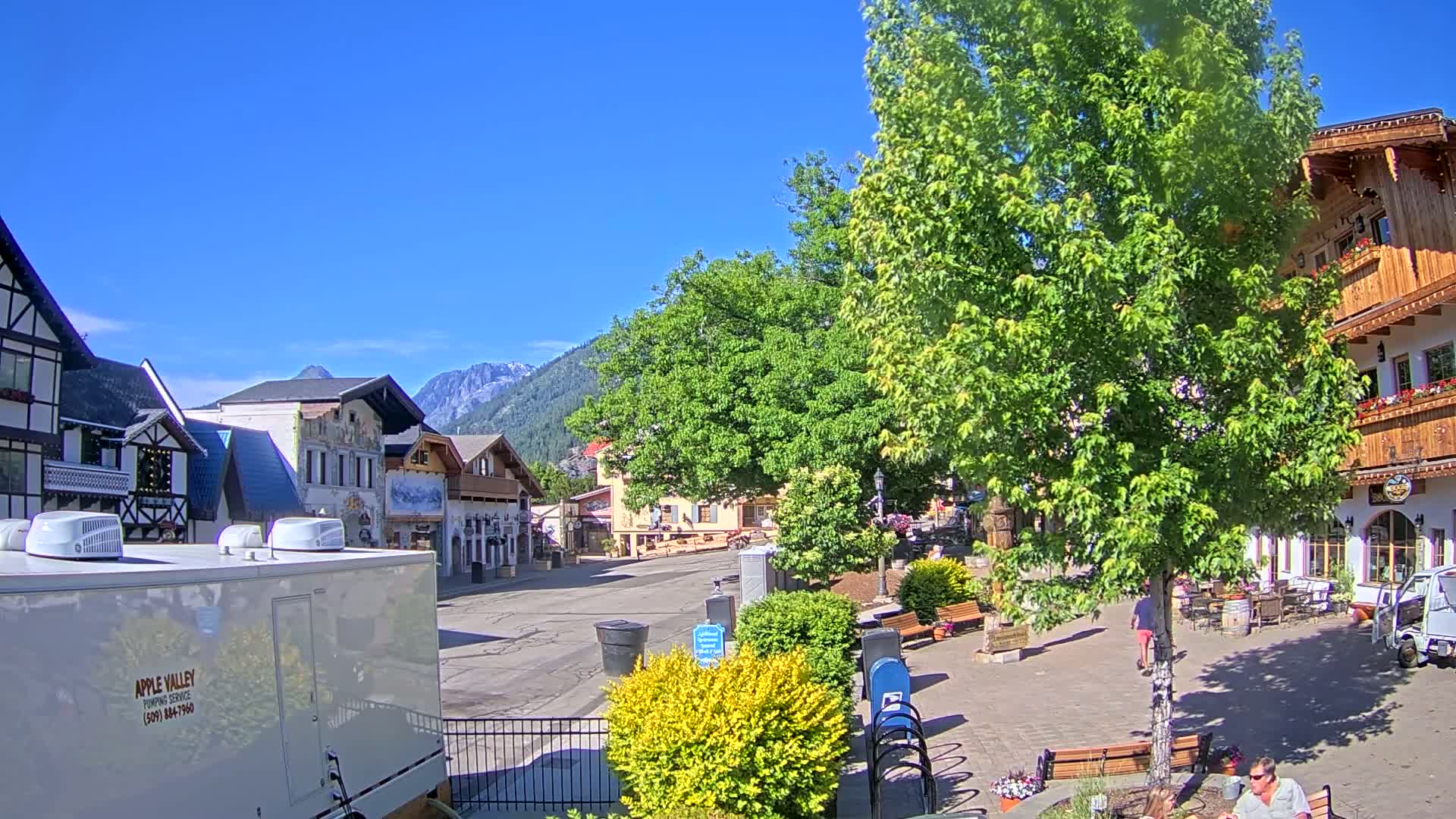 A village square with European-style buildings, lush green trees, and a clear blue sky is shown on a sunny day, with mountains in the background.