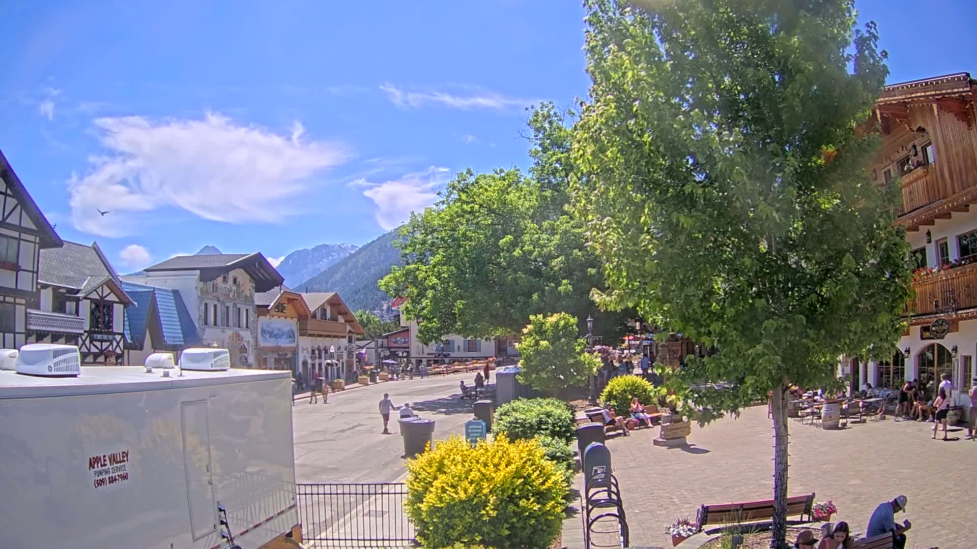 A sunny day in a Bavarian-style village square shows people relaxing and walking around under a large tree, with mountains in the background.