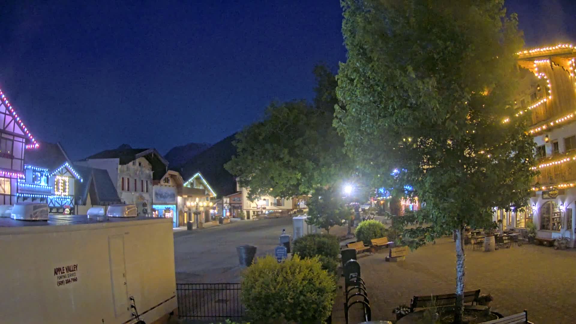 A nighttime view of a town square with several buildings decorated with string lights, under a clear, dark blue sky.