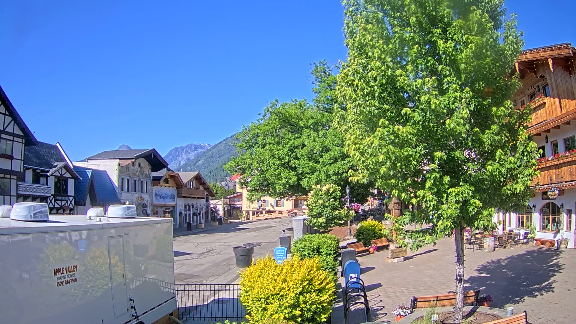 A quaint town square with European-style buildings, lush green trees, and a clear blue sky is visible under sunny conditions.