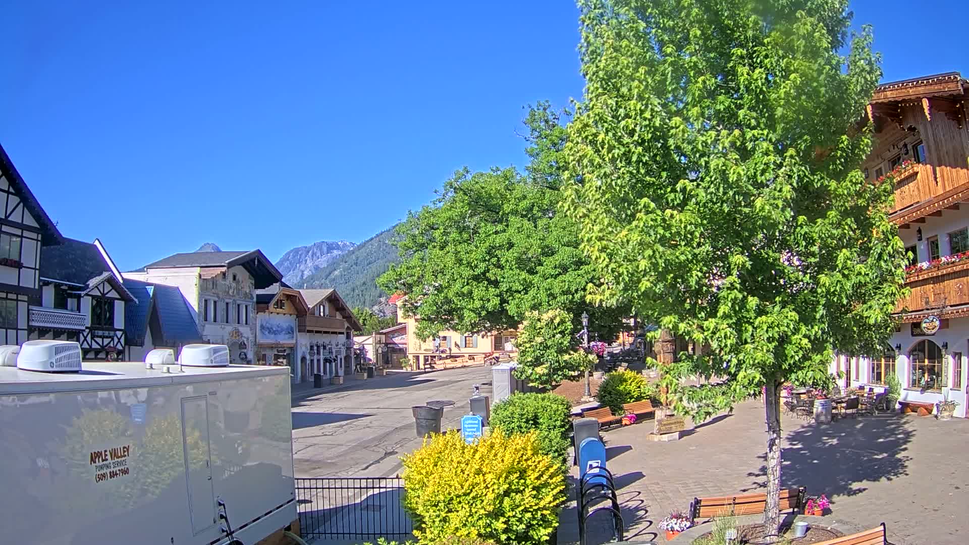 A quaint village square with European-style buildings, lush green trees, and a clear blue sky is visible on a sunny day.