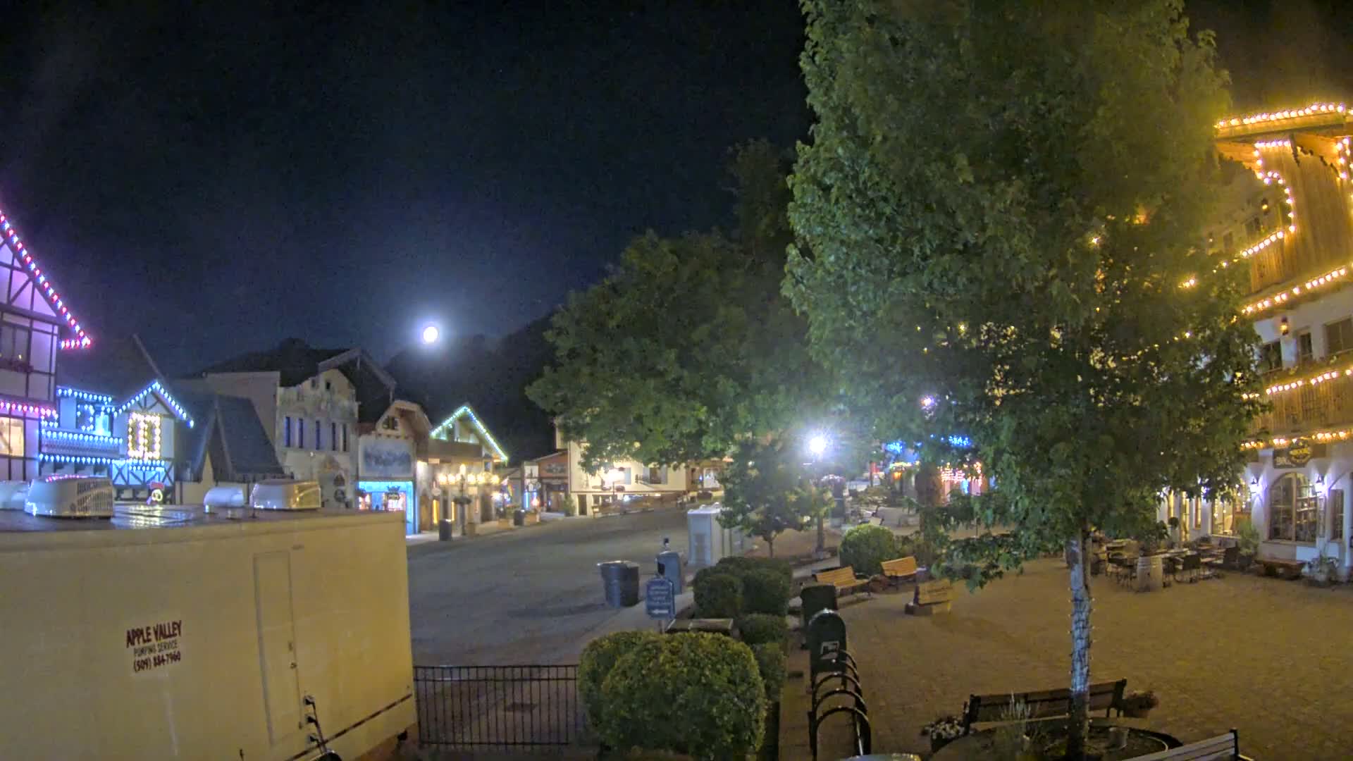 A moonlit night scene shows a town square with several buildings decorated with string lights, a large tree in the center, and a few benches.