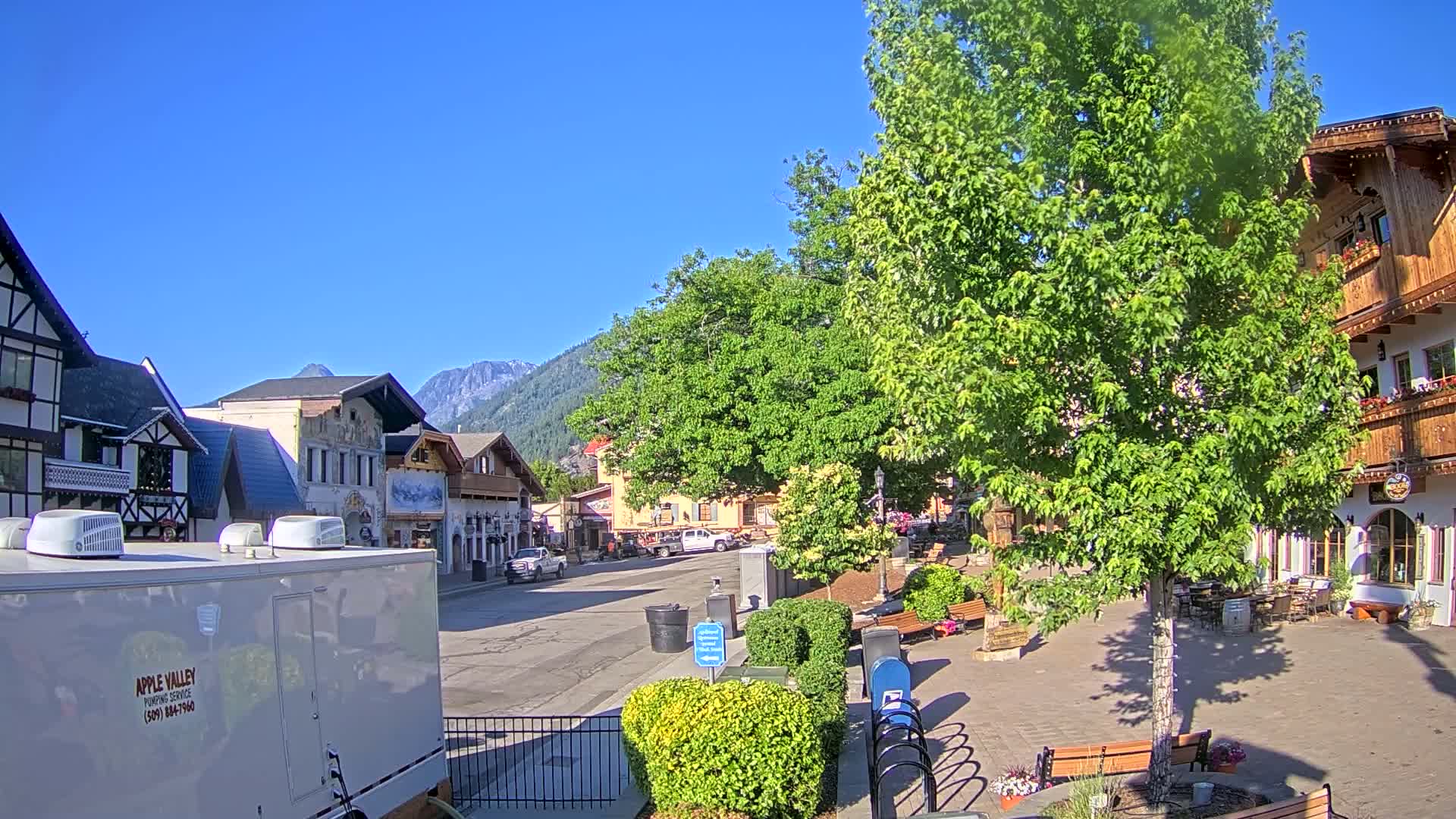 A sunny day reveals a quaint town square with European-style buildings, lush green trees, and a few parked cars, all nestled against a backdrop of mountains.