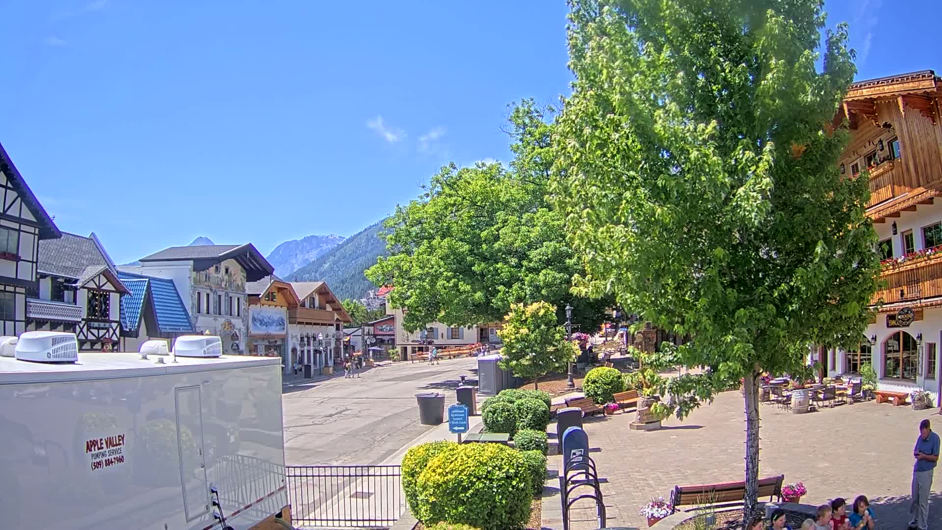 A sunny outdoor town square with European-style buildings, lush green trees, and people strolling around.