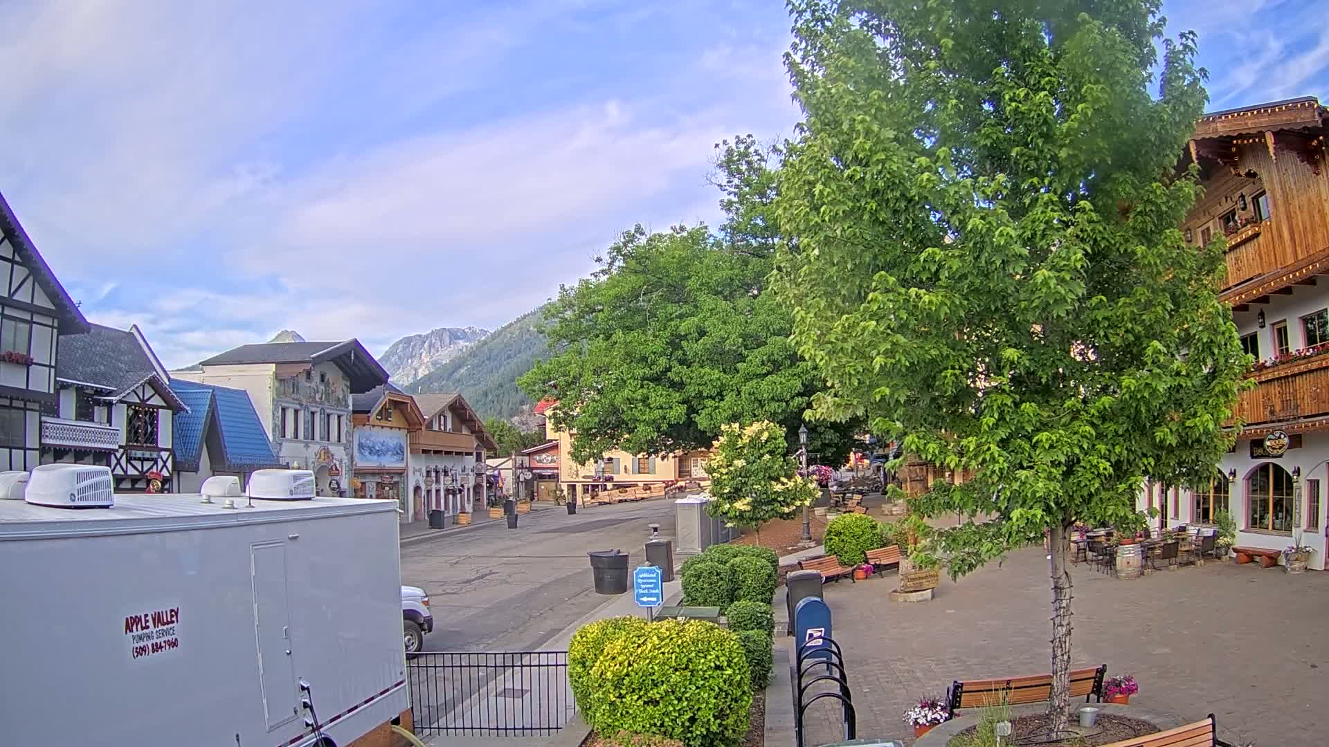 A partly cloudy day reveals a quaint town square with European-style buildings, lush green trees, and benches, nestled against a backdrop of mountains.