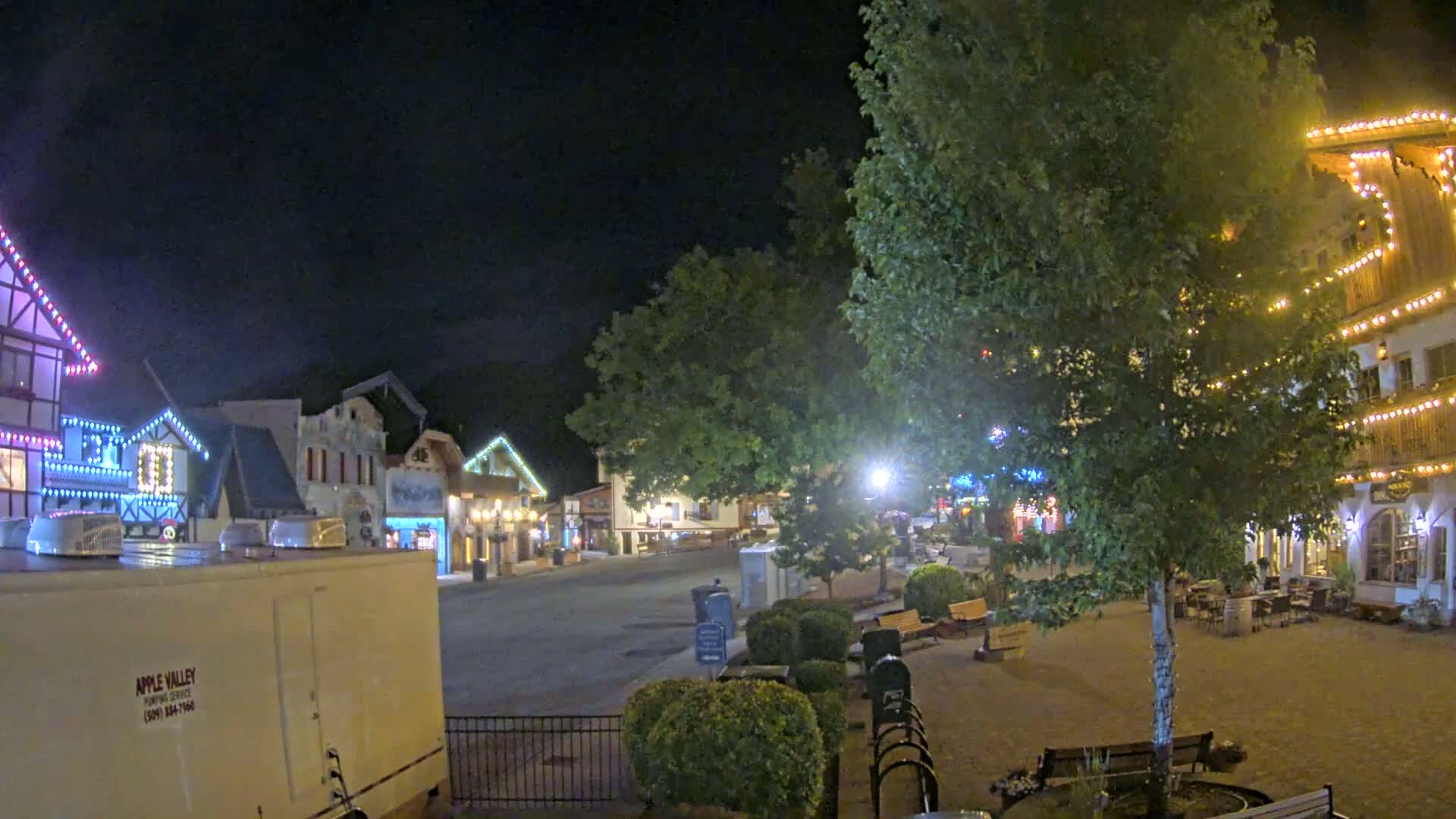 A nighttime view of a town square with several buildings decorated with string lights, a large tree in the center, and a few benches.