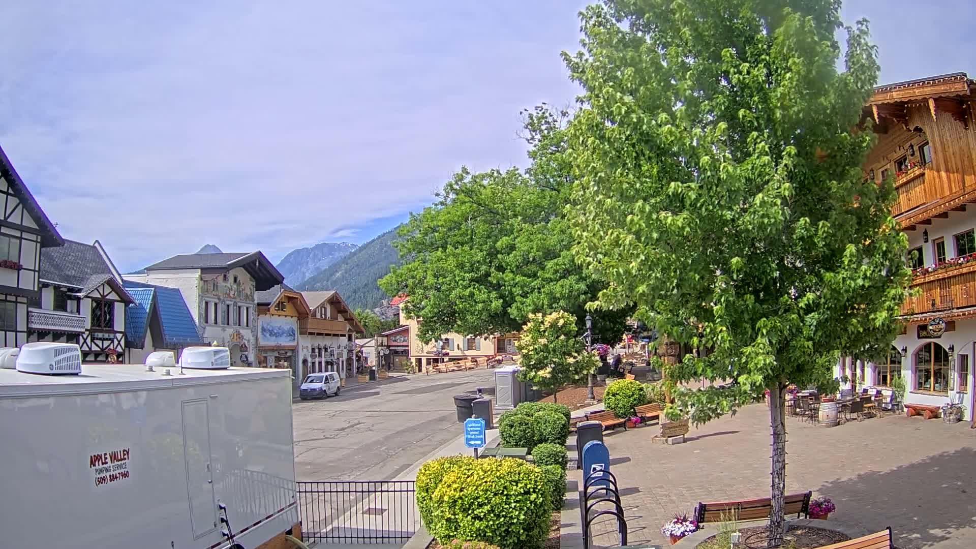 A mostly sunny day in a quaint village with European-style architecture, featuring a tree-lined street and mountains in the background.
