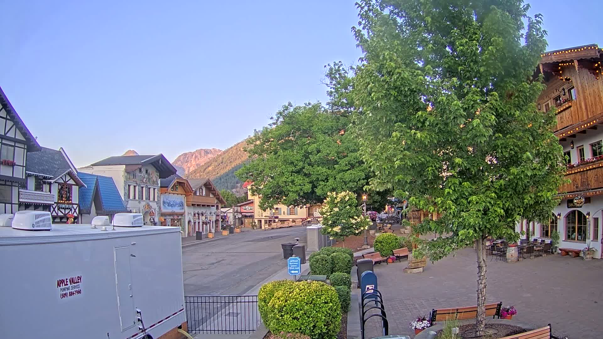 A quaint village street with European-style buildings, lush green trees, and a clear, bright sky sits at the base of a mountain range.
