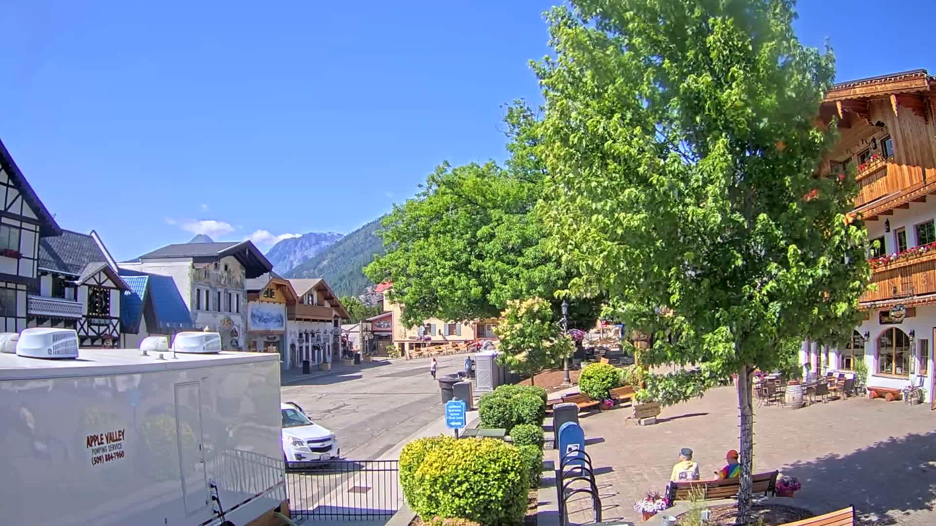 A sunny day reveals a village square with European-style buildings, lush green trees, and a few people sitting on benches, mountains in the background.