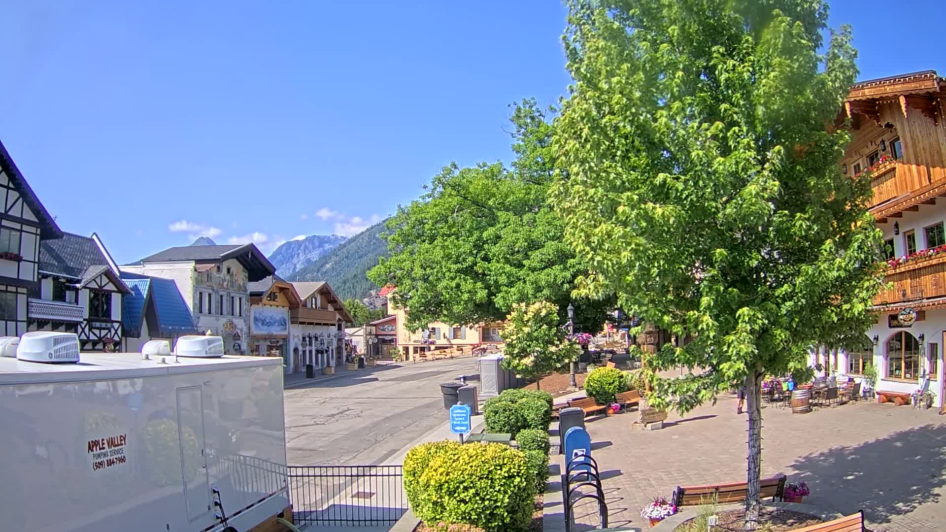 A sunny day reveals a quaint town square with European-style buildings, lush green trees, and a mountain backdrop.