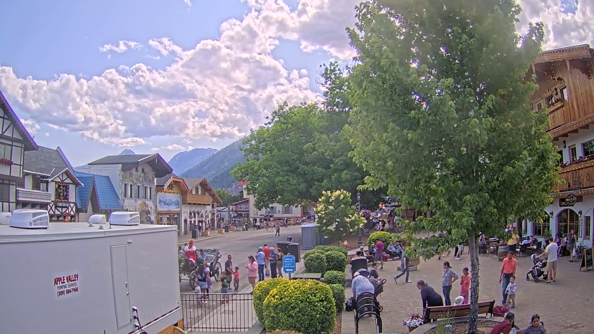 A sunny day reveals a quaint town square with European-style buildings, lush green trees, and a mountain backdrop.