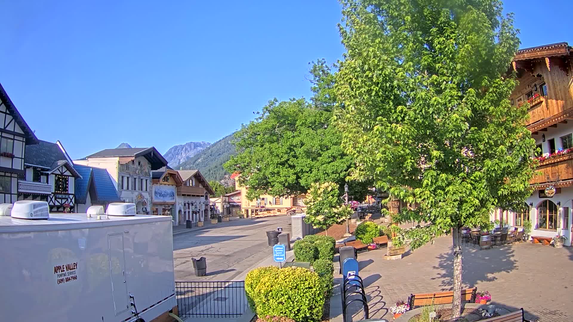 A paved town square with European-style buildings, lush green trees, and benches under a clear blue sky.