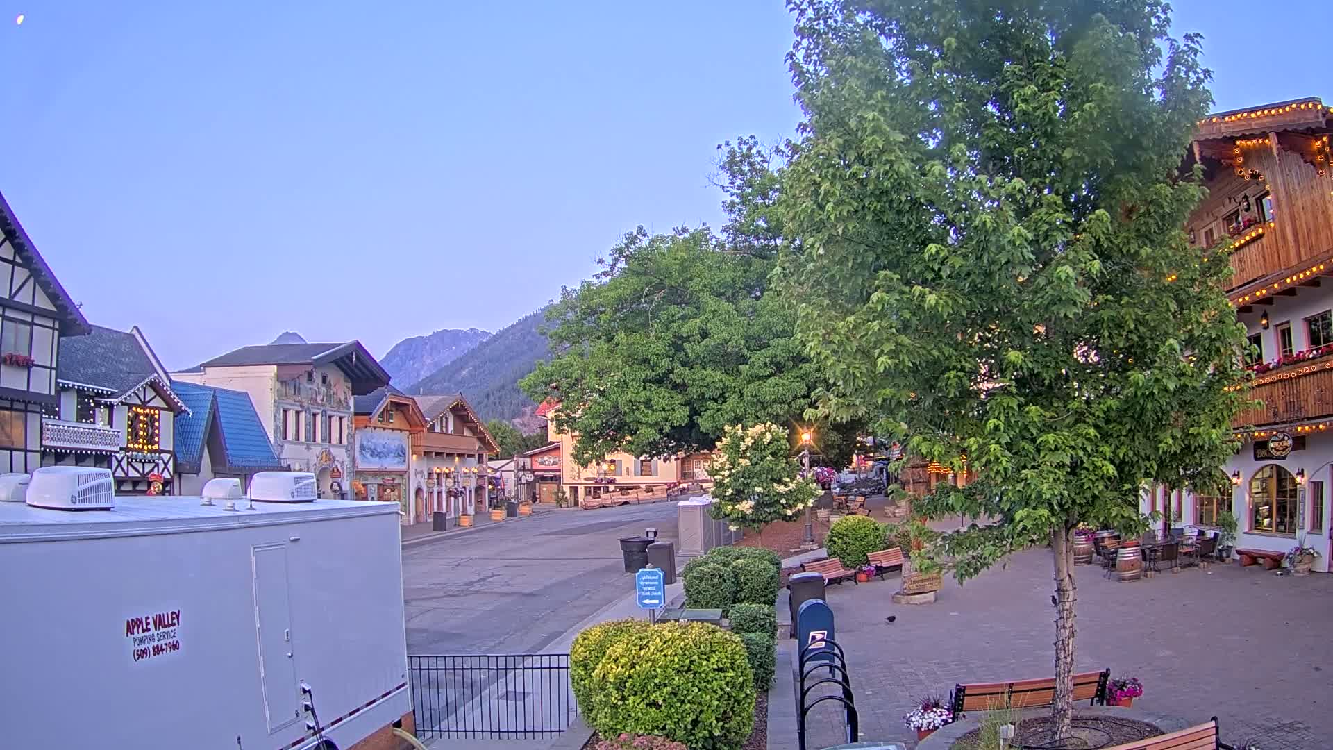 A quaint town square with European-style buildings, under a clear twilight sky, sits nestled against a mountain backdrop.