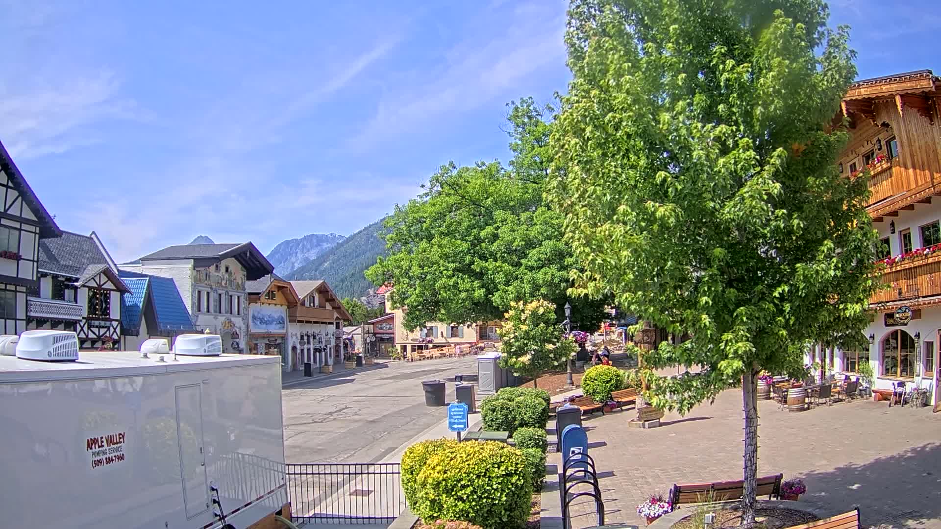 A sunny day in a Bavarian-style village with a paved town square,  lush green trees, and mountains in the background.