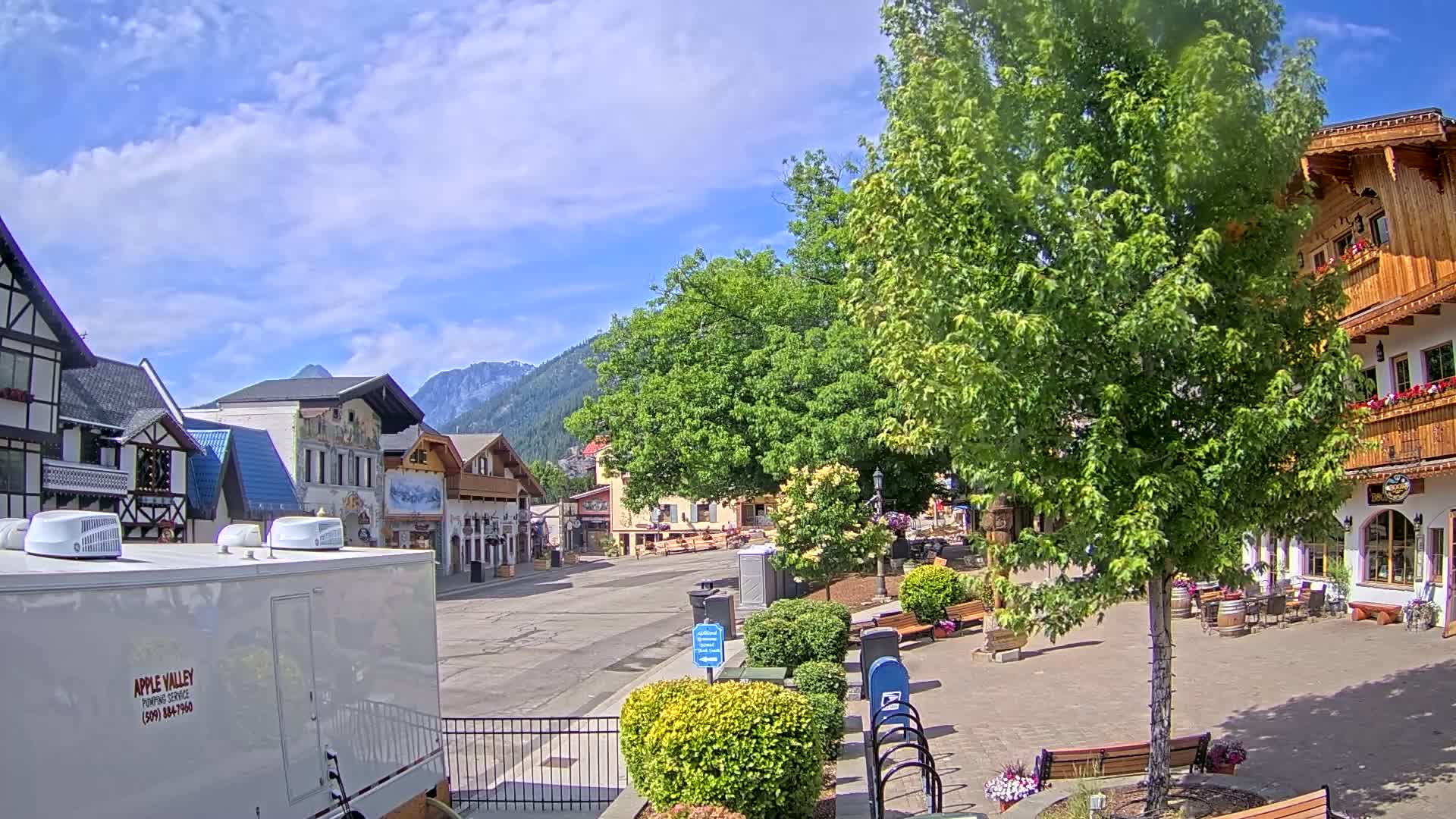 A mostly empty town square with Bavarian-style buildings, lush green trees, and mountains in the background under a partly sunny sky.