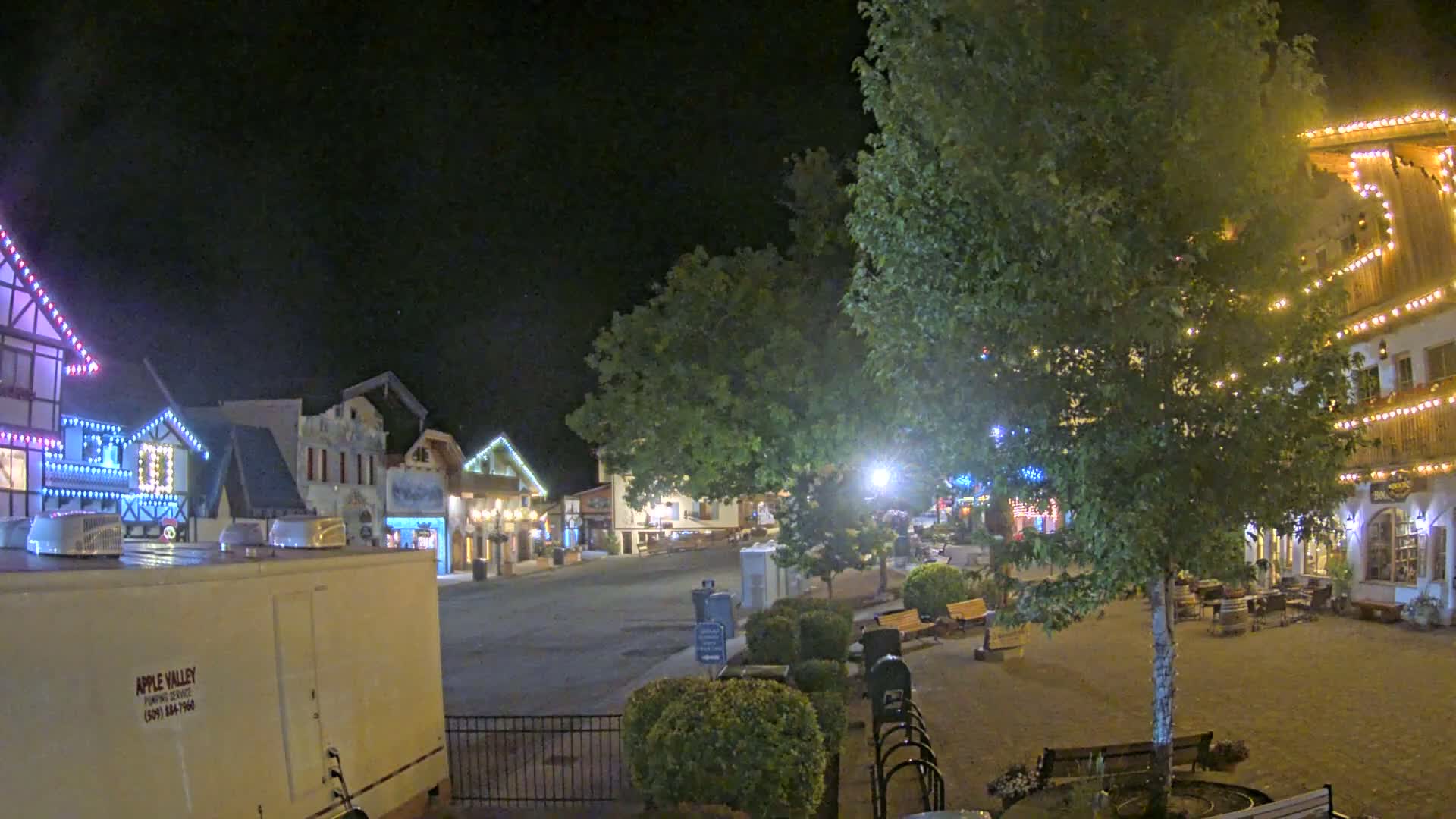 A nighttime scene of a town square with several buildings decorated with string lights, a large tree in the center, and some park benches.