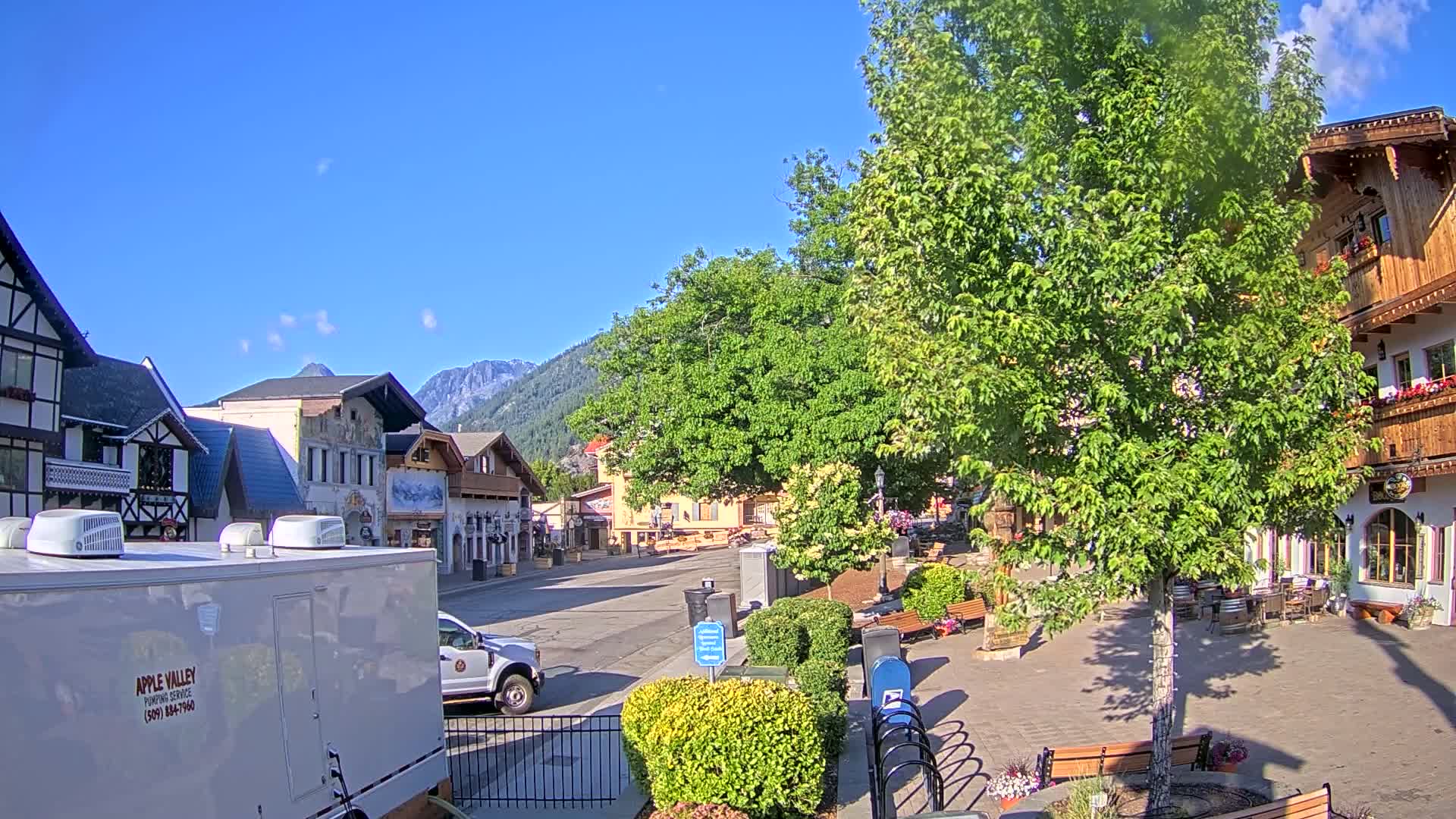 A quaint village street scene on a sunny day, with European-style buildings, lush green trees, and mountains in the background.