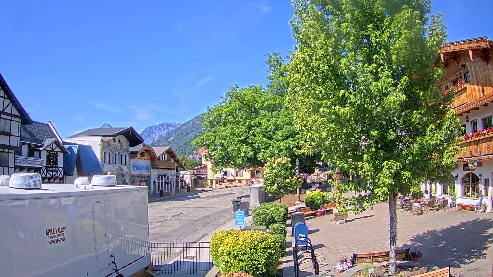 A quaint town square with European-style buildings, lush green trees, and a mountain backdrop under sunny skies.