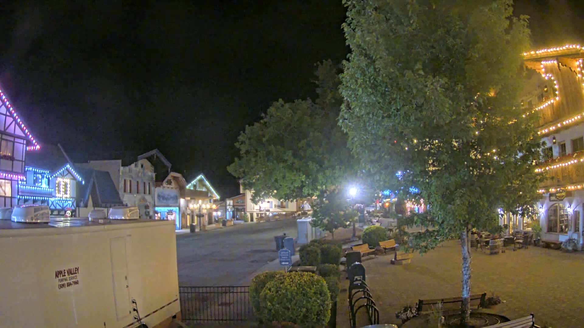 A nighttime view of a town square with several buildings decorated with string lights, a large tree in the center, and a few benches and small landscaping features.