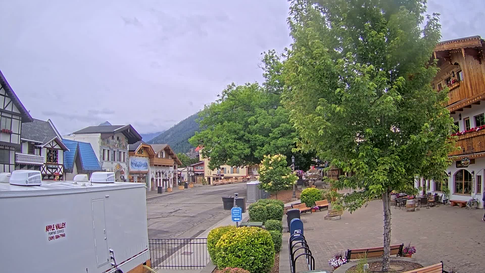 A mostly empty town square with European-style buildings, lush green trees, and mountains in the background under an overcast sky.