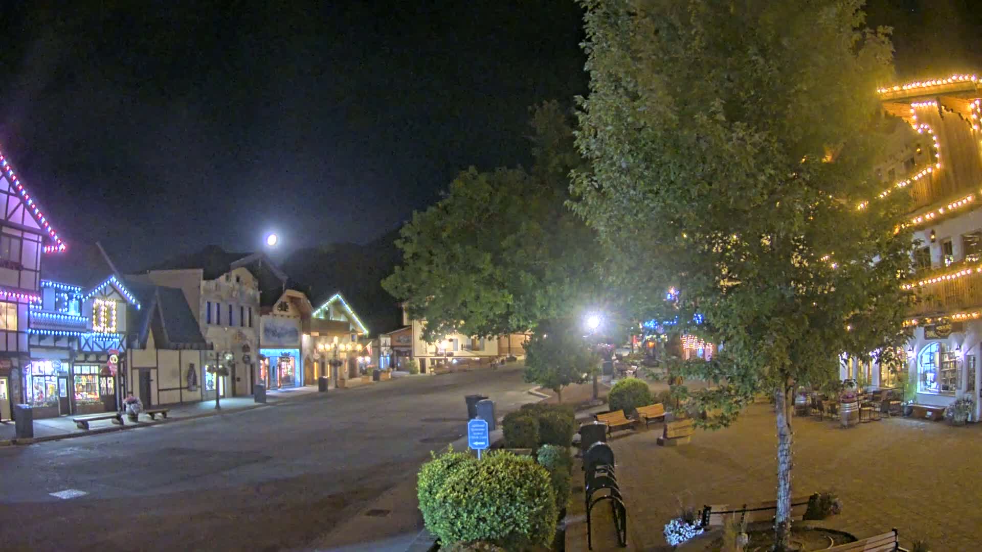 A nighttime view of a quaint town square with several buildings decorated with string lights under a full moon.