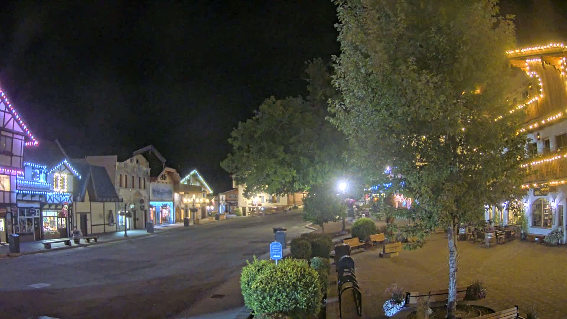A nighttime view of a quaint, festively-lit town square with several buildings adorned with string lights, a central tree, and park benches.
