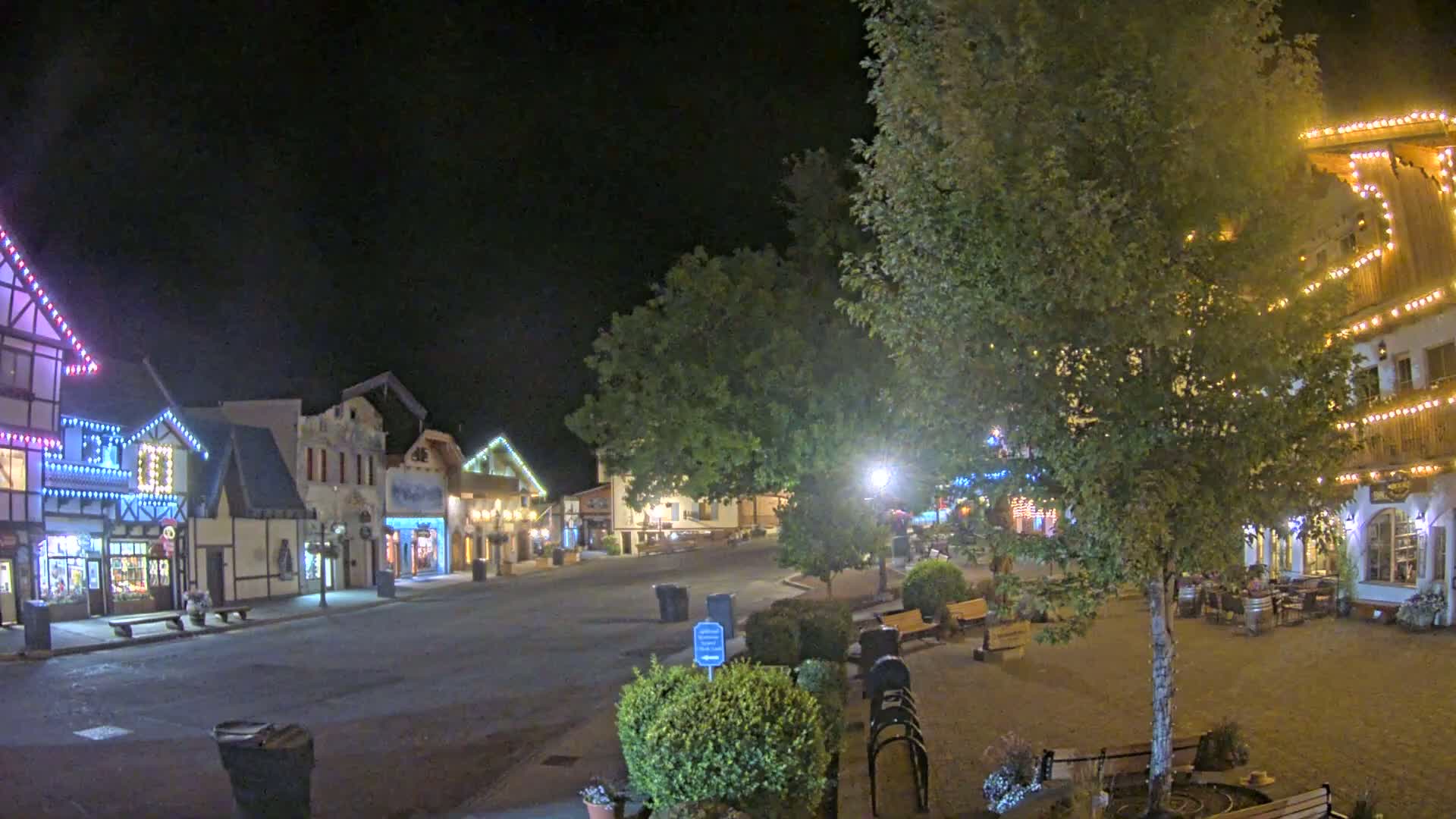 A nighttime view of a quaint, festively lit town square with several buildings, trees, and park benches.