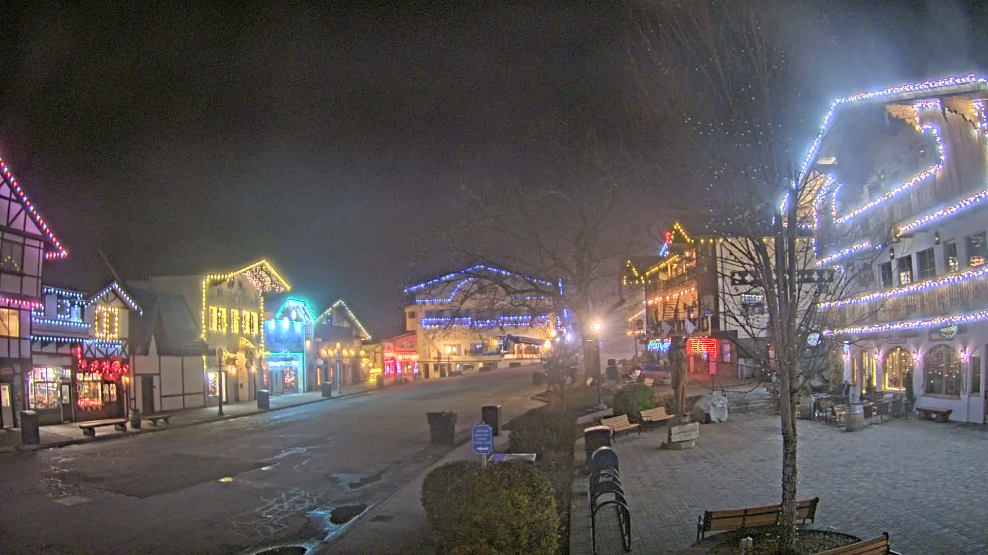 A festive European-themed village street glows with colorful string lights adorning its buildings and bare trees on a damp, dark night with wet pavement.