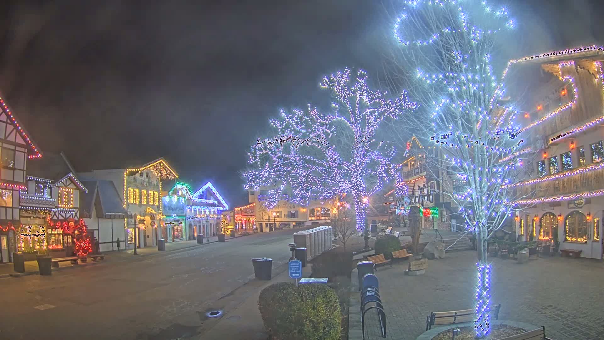 A picturesque Bavarian-style town square is brightly illuminated at night with numerous colorful holiday lights adorning buildings and trees under a clear, dark sky.