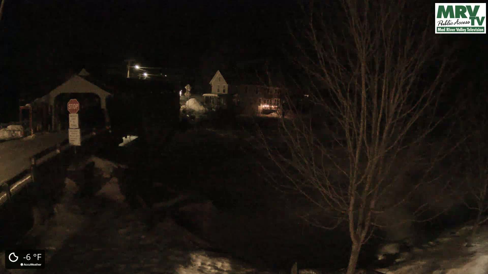 A nighttime, snow-covered scene shows a covered bridge and houses under a dark sky with a temperature of -6°F.