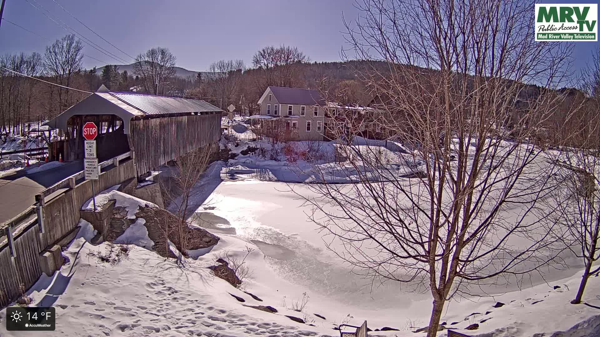 A covered bridge spans a partially frozen river under a sunny winter sky.