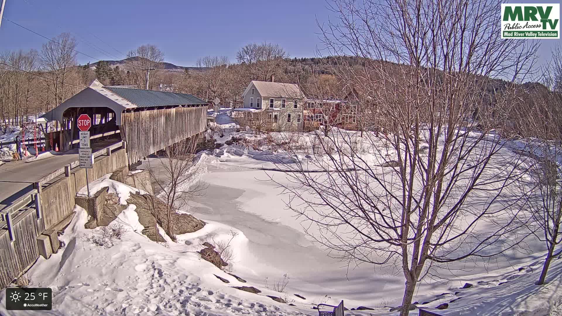 A snow-covered landscape with a wooden covered bridge, partially frozen river, and houses under a sunny sky.