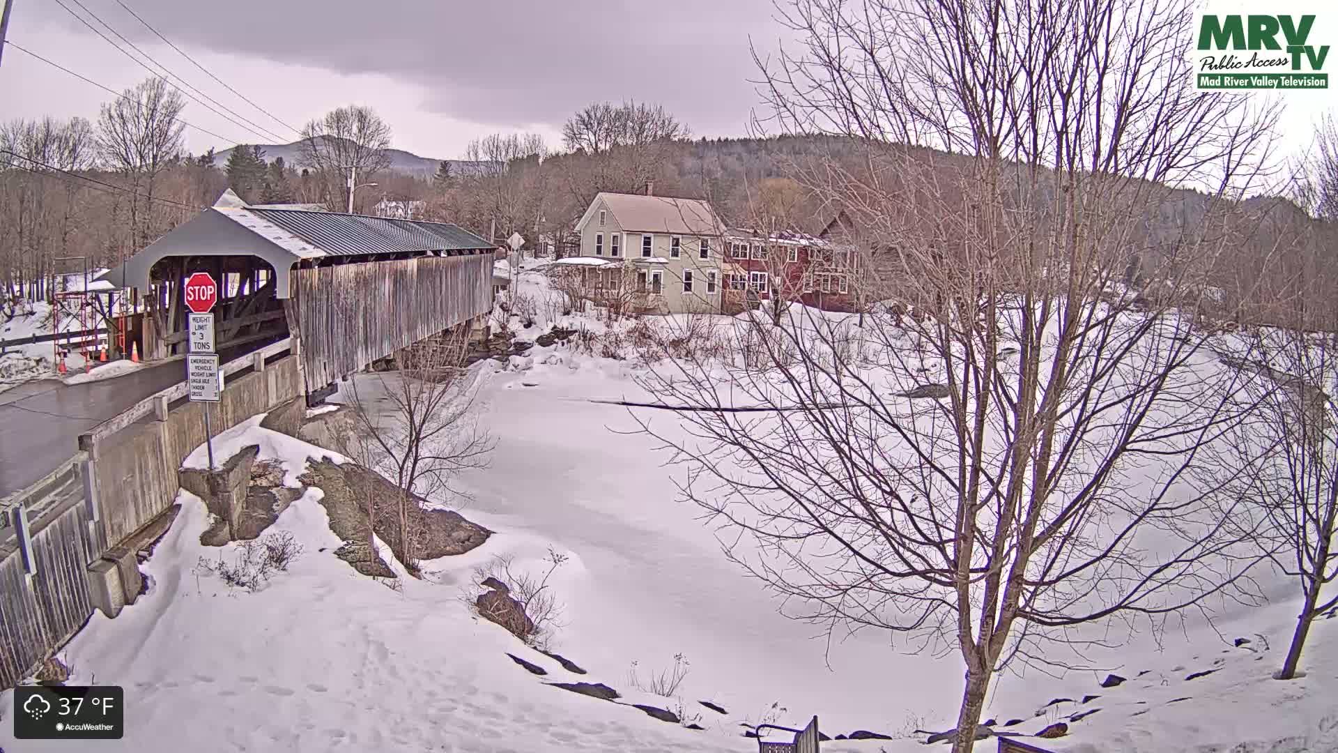 A covered bridge spans a partially frozen river in a snow-covered landscape under an overcast sky.