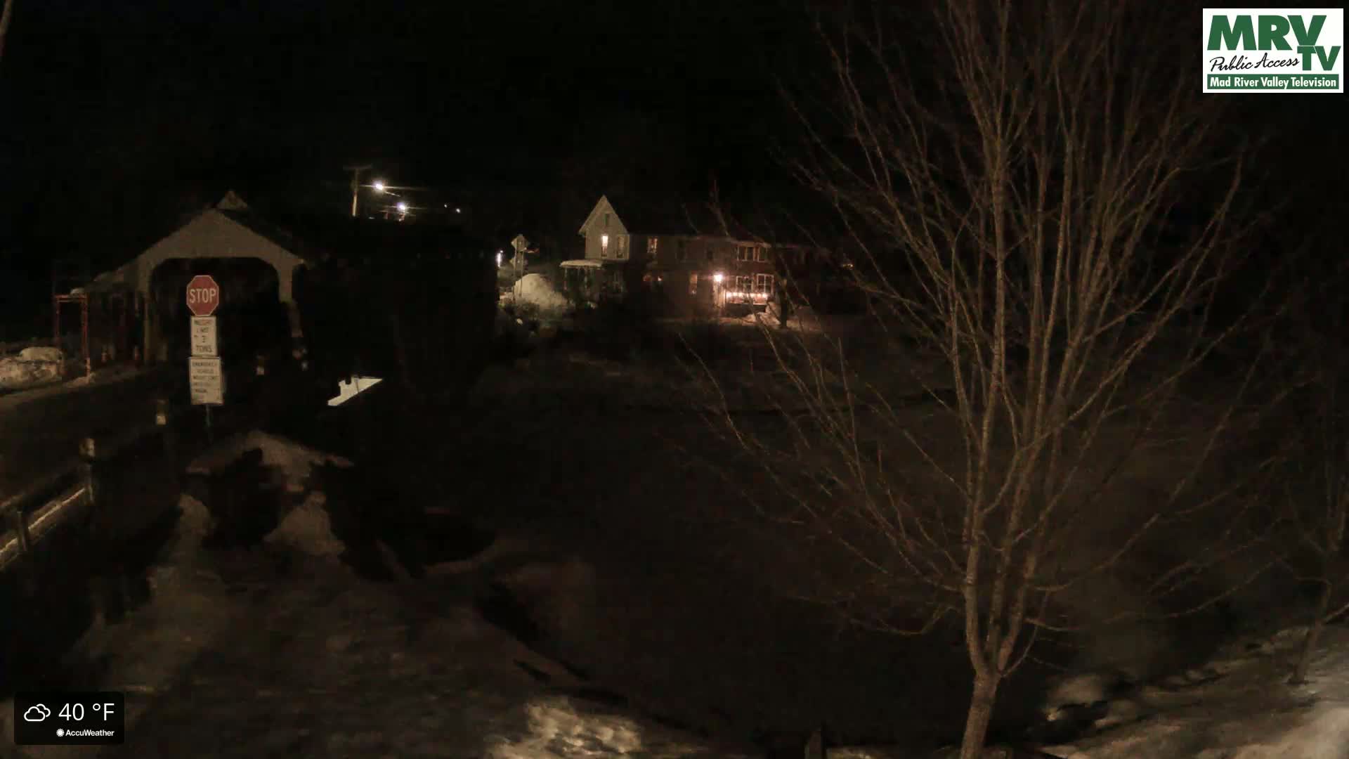 A nighttime scene shows a covered bridge, houses with lights on, and bare trees under a dark sky with snow on the ground.