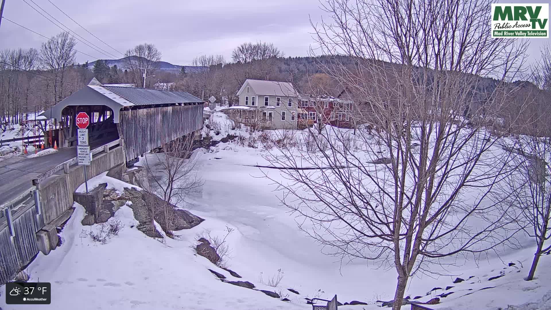 A snow-covered landscape features a covered bridge, several houses, and a partially frozen river under an overcast sky.