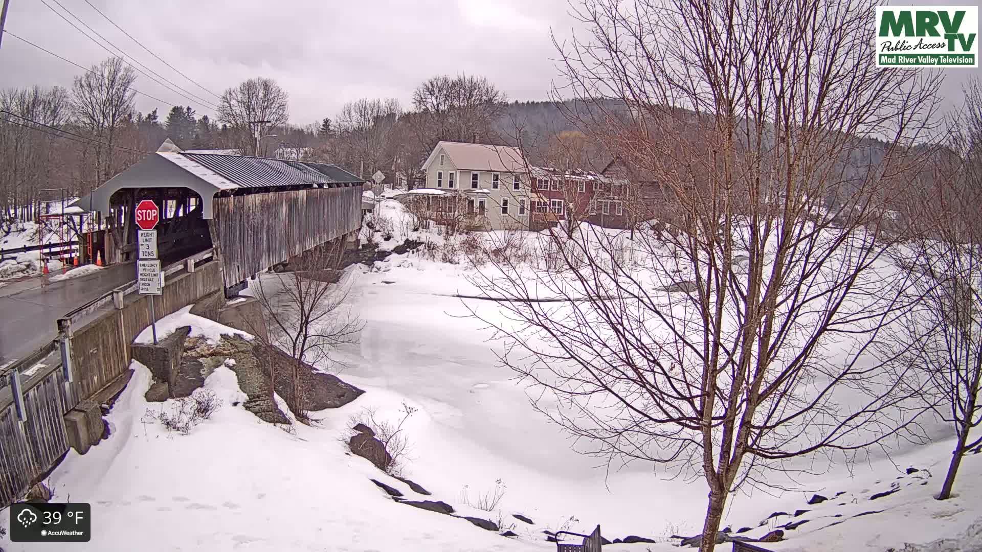 A covered bridge crosses a partially frozen river in a snowy landscape, with houses visible in the background under an overcast sky.