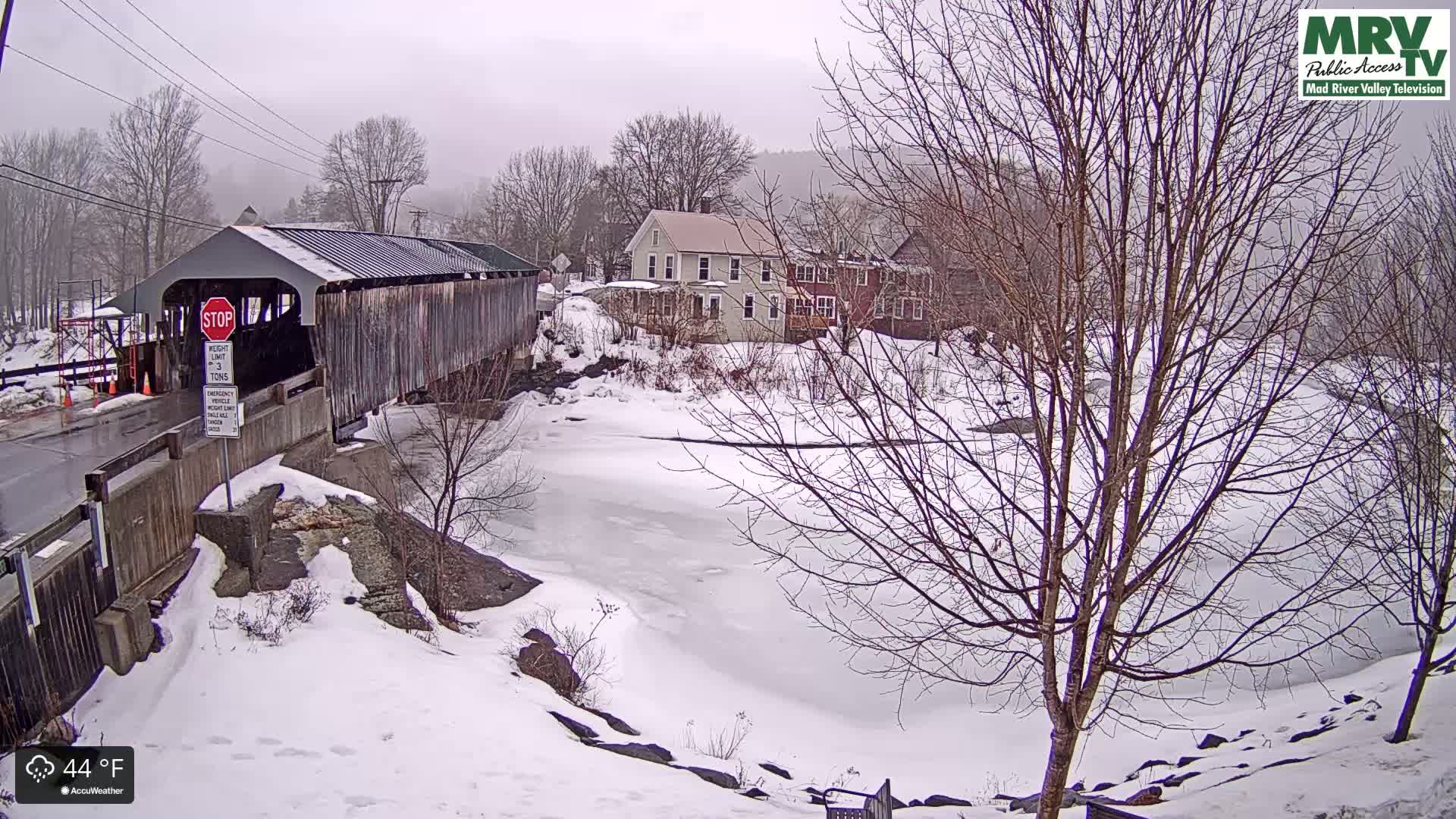 A snow-covered landscape features a covered bridge, a partially frozen river, and several buildings under an overcast sky.